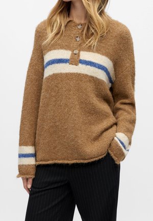 Frau trägt einen braunen, flauschigen Pullover mit weißen und blauen horizontalen Streifen und drei Knöpfen am Kragen, kombiniert mit schwarzen Nadelstreifenhosen.