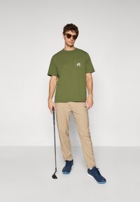 Olive green cotton t-shirt with a small white pocket logo, paired with beige pants and navy sneakers. Holding a golf club.