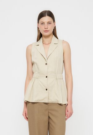Mujer con chaleco beige sin mangas con botones y cintura con peplum, y pantalones marrones, de pie frente a un fondo blanco liso.