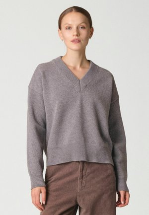 Femme portant un pull gris ample à col en V et un pantalon en velours côtelé marron, debout devant un fond clair uni.