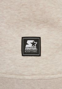 Tissu beige avec un patch carré noir présentant un logo blanc stylisé en forme de "S" et une étoile au-dessus des mots "Starter Black Label".
