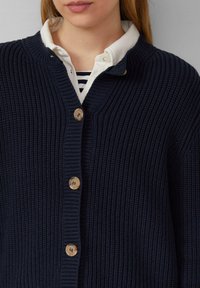 Dunkelblau gerippter Cardigan mit V-Ausschnitt, ausgestattet mit Holzknöpfen und einem strukturierten Garn. Getragen über einem weißen, gestreiften Hemd mit Kragen.
