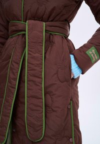 Brązowy pikowany szlafrok z zielonymi lamówkami i paskiem, ozdobiony stębnowaniem w kształcie jabłek. Zawiera kieszeń oraz naszywkę z tekstem na rękawie.