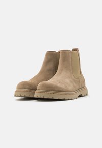 Botine din zamac, de culoare bej deschis, cu panouri laterale elastice, vârful rotund și tălpi din cauciuc texturat, având un model de profil pentru aderență.