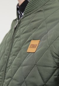 Jachetă bomber matlasată verde, cu guler cu nervuri și un ecuson din cauciuc pe pieptul stâng, de culoare bej. Textură netedă și design ajustat.