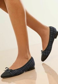 Marineblaue Ballettschuhe mit einem strukturierten Muster und schwarzem Zehenkappen, getragen von Beinen mit einem dezenten Glanz, vor einem hellen Hintergrund.