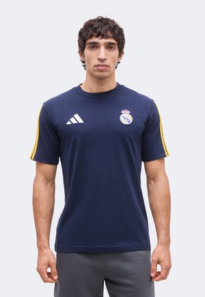 Bărbat purtând un tricou albastru închis Real Madrid Adidas cu dungi galbene pe mâneci, stând pe un fundal simplu gri deschis.