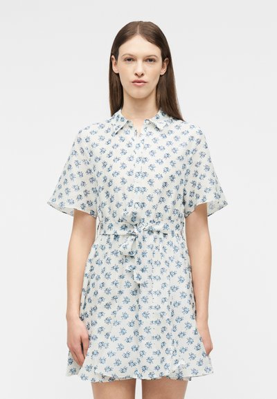 Femme portant une robe chemise blanche à manches courtes avec des imprimés floraux bleus et une ceinture nouée à la taille, debout devant un fond blanc uni.