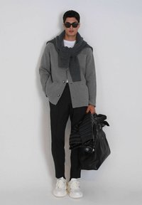 Grijze textuurjacket met zakken, gelaagd over een kledingstuk met ronde hals. Zwarte broek, witte sneakers, en houdt een zwarte pufferjack en tas vast.