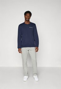 Felpa blu navy con scollo a girocollo e logo bianco sul petto, abbinata a pantaloni della tuta grigio chiaro, indossata con scarpe da ginnastica bianche.