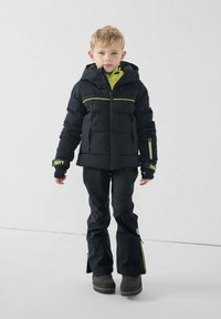 Chaqueta aislante negra con detalles en verde, combinada con pantalones impermeables negros. Material texturizado, diseño con capucha, puños en capas y cremalleras laterales.