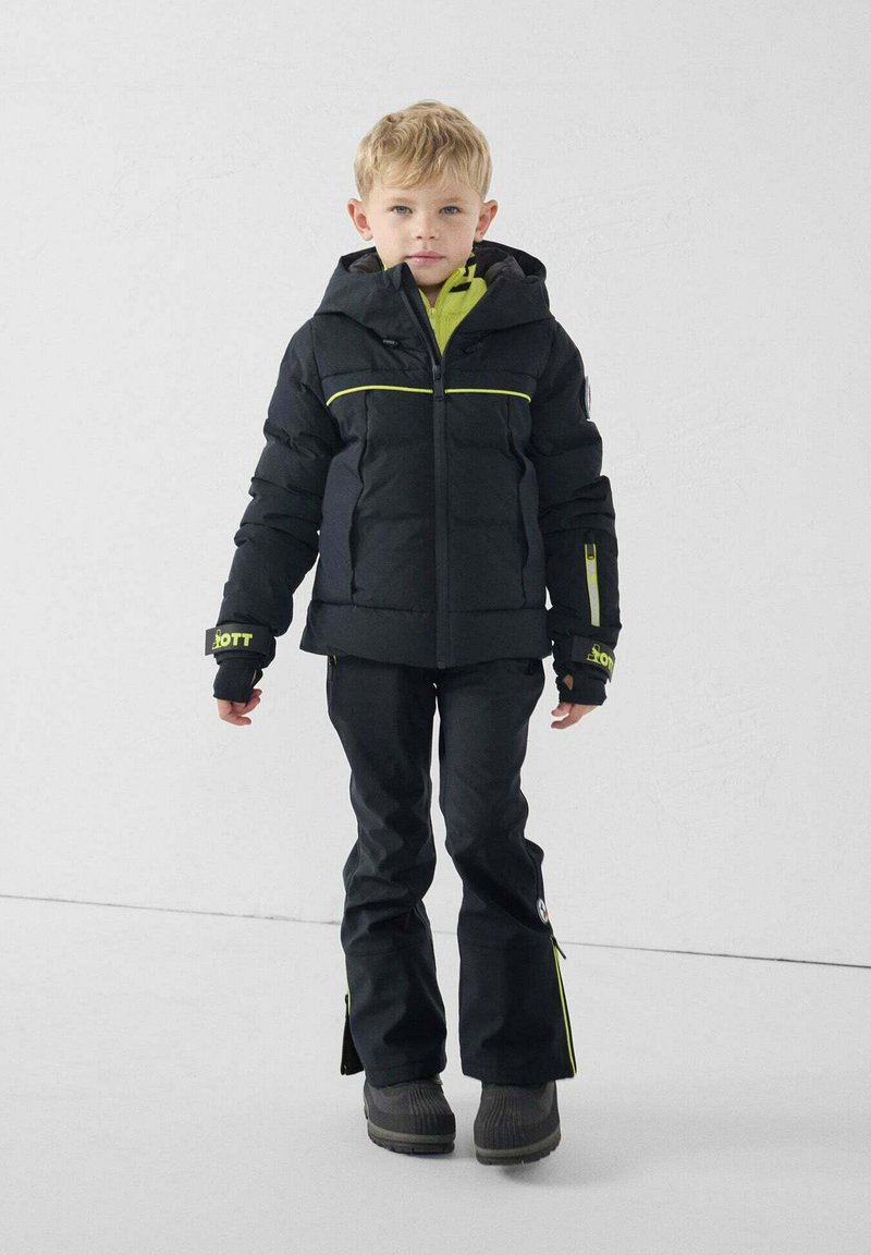 Chaqueta aislante negra con detalles en verde, combinada con pantalones impermeables negros. Material texturizado, diseño con capucha, puños en capas y cremalleras laterales.