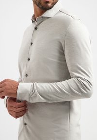 Helle graue Langarmbluse aus glattem Stoff mit Knopfleiste, spitzem Kragen und schwarzen Knöpfen. Eng anliegendes Design um die Arme.