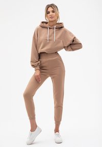 Beżowy hoodie z białymi sznurkami i kieszenią kangurką, zestawiony z beżowymi dopasowanymi dresami. Model nosi białe sneakersy. Gładka tekstura tkaniny.