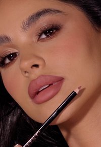 Matowy nagi szminka i konturówka trzymane blisko ust. Gładkie wykończenie, subtelny połysk na powiekach, wyraźne rzęsy, naturalny odcień skóry i jasne brwi.