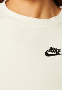 Närbild av en vit T-shirt med rund halsringning och en svart Nike-logotyp broderad på övre bröstet.