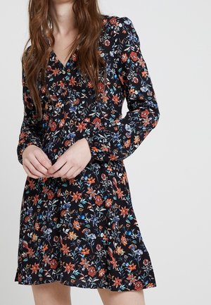Femme portant une robe noire à manches longues avec des fleurs rouges, orange et bleues, debout, les mains jointes devant elle.