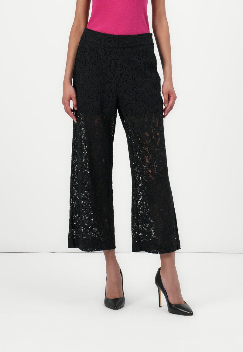 Pantalons culottes en dentelle noire avec une jambe large, dotés d'un ourlet à volants et d'un motif floral complexe. Portés avec des chaussures à talons hauts noires.