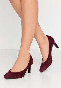 Ben gömd med korslagda ben i burgundy mockapumps med medelhögt klack, som står på en vit yta med en neutral ljus bakgrund.