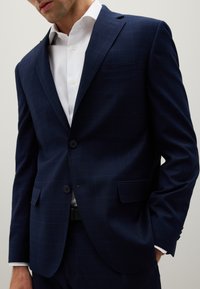 Giacca da abito a quadri blu navy con rever a taglio, due bottoni e taschino sul petto; indossata sopra una camicia bianca con bottoni visibili.
