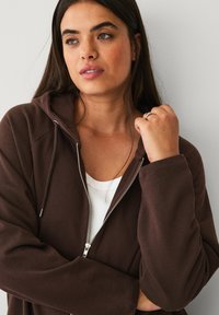 Brown zip-up hoodie made of soft fabric, featuring a hood, front pockets, and silver hardware, layered over a white tank top.