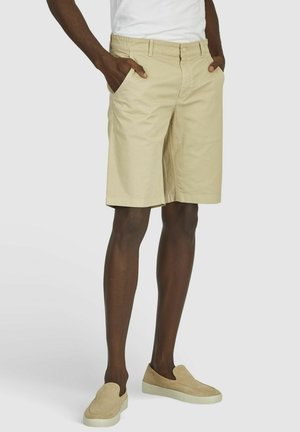 Beige Baumwollshorts mit Seitentaschen, geradem Bein und Knopfverschluss. Kombiniert mit hellbeigen Slippers.