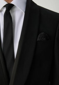 Schwarze Sakkojacke mit satiniertem Revers, weißes Hemd, schwarze Krawatte und ein gemusterter schwarzer Einstecktuch in der Brusttasche.