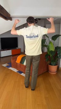 Moški, ki stoji v dnevni sobi z dvignjenimi rokami, oblečen v kremno Timberland majico in olivno zelene hlače, zraven lončnice in kavča.