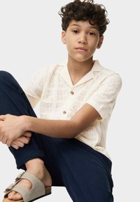 Camisa polo de manga corta color crema con un patrón de tejido texturizado y tres botones en la parte frontal. Combinada con pantalones azul marino y sandalias gris claro.