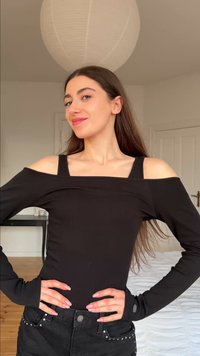 Νεαρή γυναίκα με μακριά καστανά μαλλιά φοράει μαύρο τοπ ώμου και μαύρα τζιν με καρφιά, στέκεται με τα χέρια στη μέση σε φωτεινό δωμάτιο.