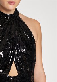 Crni halter top sa šljokicama, visokim ovratnikom, prorezom u obliku ključanice i teksturiranim dizajnom, s prilagođenim siluetom i sjajnim detaljima.