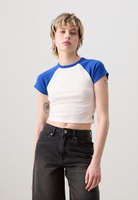 Top corto in cotone bianco con maniche corte raglan blu. Il modello lo indossa con jeans neri a vita alta, mettendo in mostra una silhouette aderente.