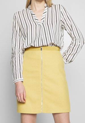 Femme portant un chemisier blanc à rayures verticales noires et une jupe jaune taille haute avec une fermeture éclair à l’avant.