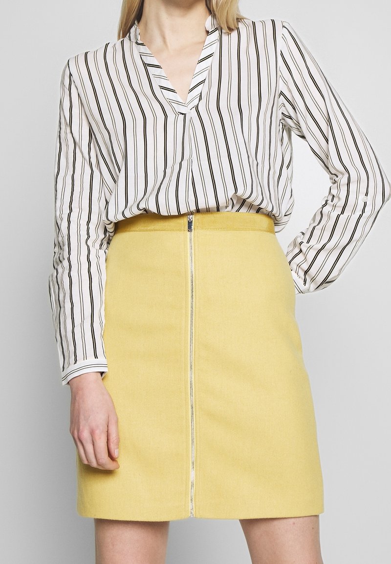 Femme portant un chemisier blanc à rayures verticales noires et une jupe jaune taille haute avec une fermeture éclair à l’avant.