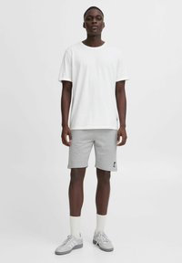 T-shirt en coton blanc, shorts en coton gris avec logo noir, baskets grises et chaussettes blanches aux chevilles. Tenue athlétique simple et décontractée.