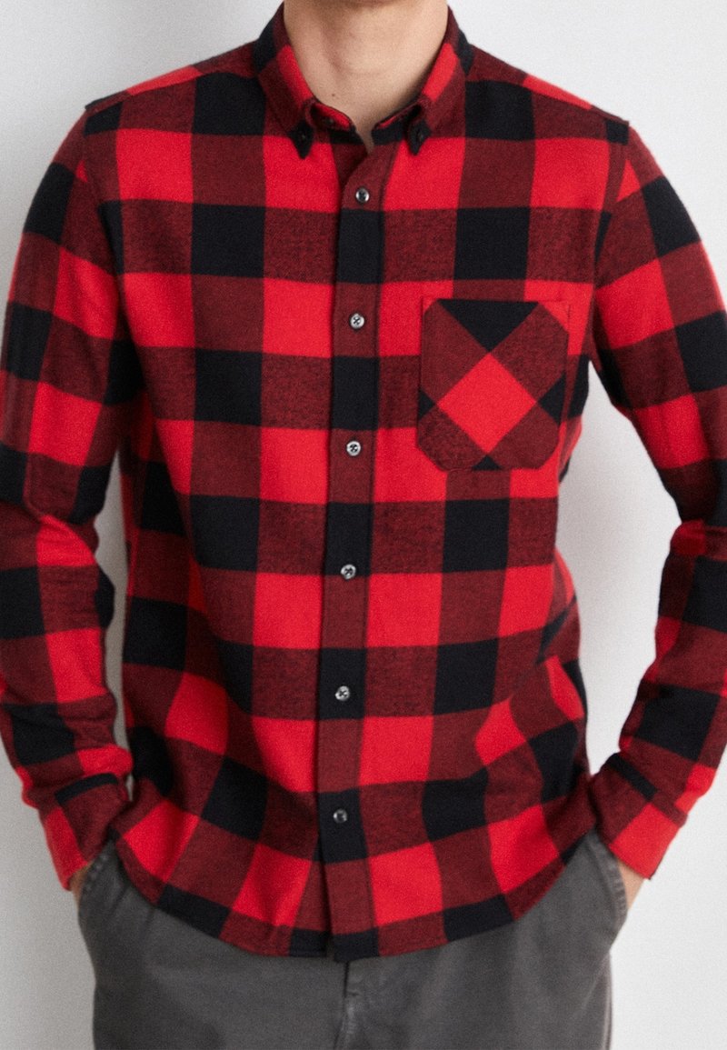 Camisa de franela a cuadros roja y negra con cuello abotonado, bolsillo frontal y botones negros. Mangas largas con puños abotonados.