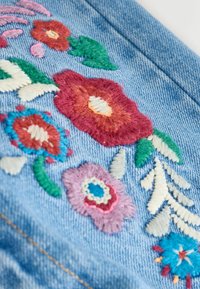 Denimstoff mit buntem Blumenstickereien. Enthält pinke, rote und blaue Blumen, grüne Blätter und cremefarbene Akzente auf einem hellblauen Hintergrund.