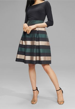 Nainen pukeutuneena mustaan neljänneshihaiseen toppiin ja laskostettuun hameeseen, jossa leveät vaakasuorat metallinhohtoiset kullan, vihreän ja mustan raidat, yhdistettynä mustiin nilkkaremmi-korkokenkiin.