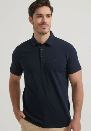 Jeune homme portant un polo bleu marine à manches courtes et un pantalon beige, debout avec une main dans la poche, fond neutre.