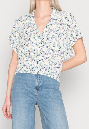 Femme portant un chemisier cache-cœur à manches courtes à fleurs avec col en V et jean bleu taille haute sur un fond uni.
