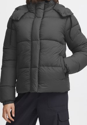 Vrouw draagt een zwarte doorgestikte gewatteerde jas met hoge kraag en hand in de zak, gecombineerd met zwarte cargobroek.