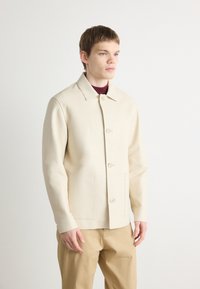 Beige jacket made of smooth fabric, featuring a collar and six buttons, with two front pockets and a tailored fit. Paired with light khaki trousers.