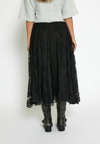 Gonna midi in pizzo nero con motivi intricati, caratterizzata da una finitura testurizzata e un design a strati. Abbinata a stivali alti fino al ginocchio con tacco spesso.