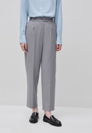 Personne portant un pantalon gris taille haute à plis et des mocassins noirs, associé à une chemise bleu clair boutonnée rentrée dans le pantalon.
