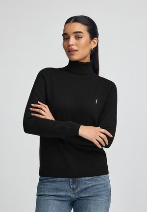 Vrouw met donker haar in een zwarte coltrui en blauwe jeans, met gekruiste armen tegen een egale achtergrond.