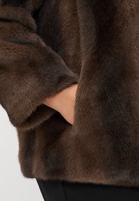 Brązowa kurtka z sztucznego futra o miękkiej fakturze i luźnym kroju. Rękaw posiada ukrytą kieszeń, co zwiększa funkcjonalność i styl.