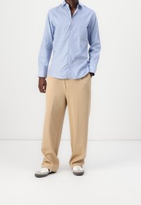 Hombre vestido con camisa de cuadros azul claro, pantalones de pierna ancha beige y zapatillas blancas y grises, de pie con una mano en el bolsillo.