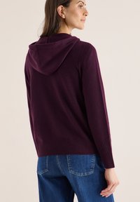 Sudadera con capucha en burdeos hecha de una tela suave, con puños y dobladillo acanalados, combinada con jeans azules. Detalle de textura sutil en la tela.