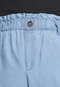 Světle modré kalhoty s elastickým pasem a jedním hnědým knoflíkem, vyznačující se jemnou strukturou a uvolněným střihem.