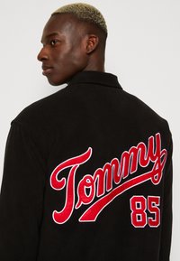 Giaccone in pile nero con grande scritta ricamata rossa "Tommy" e numero "85" sul retro. Collo a punta e texture liscia.